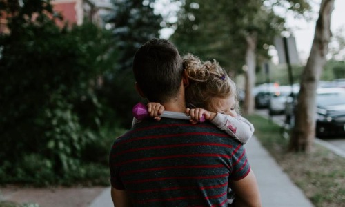 Close to the center of everything tired little girl rests her head on her father's shoulder as he carries her down the city sidewalk